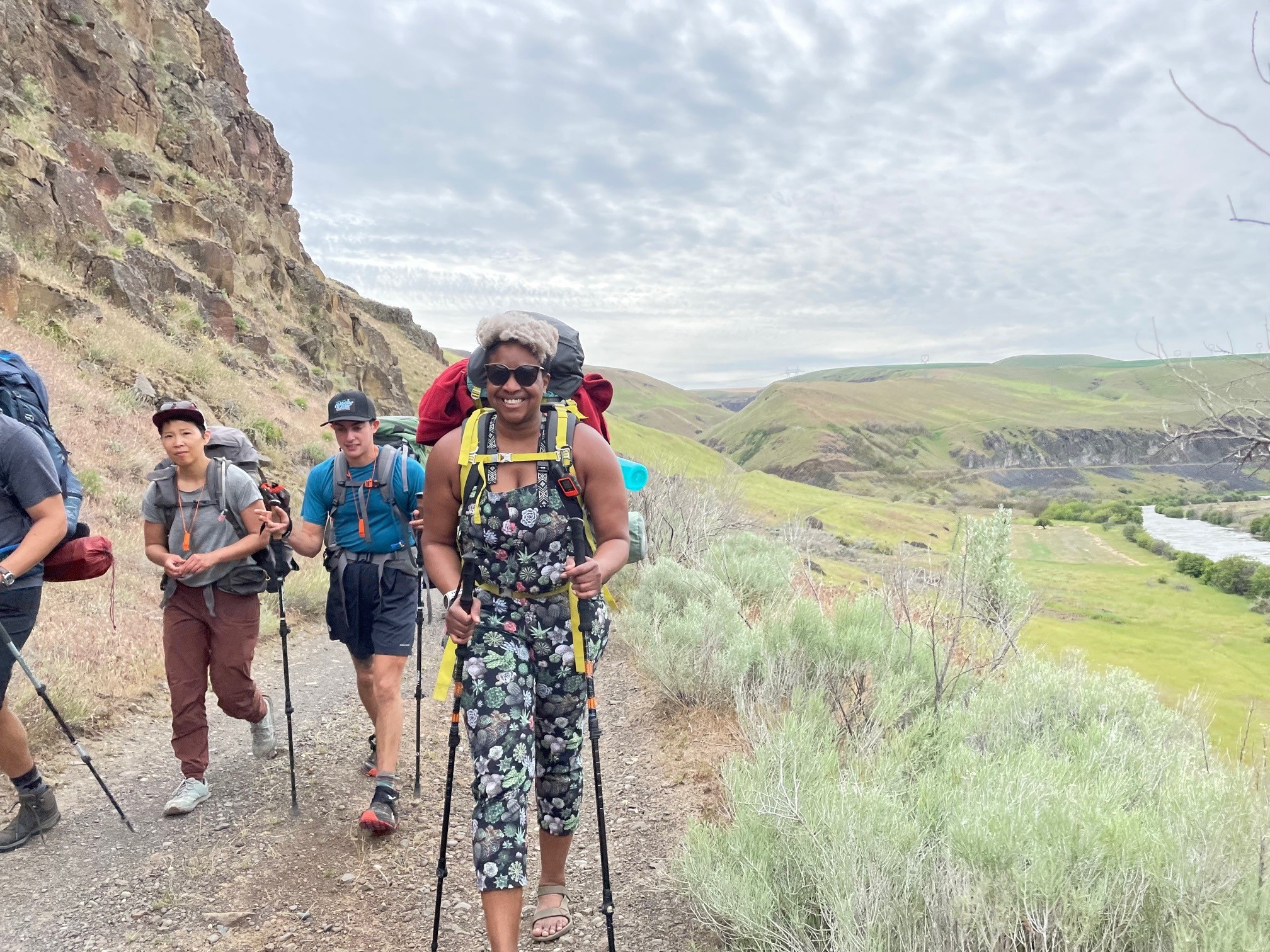A group of people walk down a trail with backpacks on. The person in the center smiles at the camera.